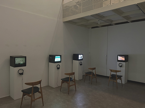 Photo of a dimly lit exhibition space. Four small, old-fashioned monitors with headphones are standing on plinths along two walls. In front of each a wooden chair.