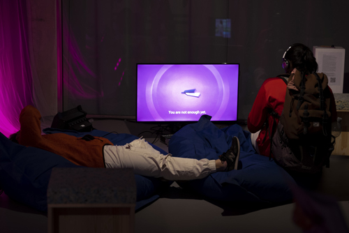 Two people with headphones sitting and laying in front of a video monitor. On the monitor a 3D model of a tuning fork is surrounded by a circle and captions that read: “You are not enough yet.” 
